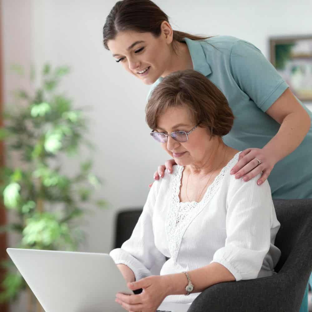 a young caregiver supporting an elderly woman working on a laptop, symbolizing guidance and assistance in a warm, comfortable environment.